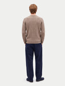 A man viewed from behind wearing a beige cable-knit sweater and navy blue trousers, standing against a light gray studio background.