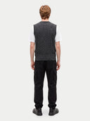 A man viewed from the back wearing a dark gray cable-knit sweater vest over a white t-shirt, black cargo pants, and black shoes, against a light gray studio background.