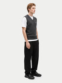 A man wearing a dark gray cable-knit sweater vest over a white t-shirt, paired with black trousers and black shoes, standing against a light gray studio background.