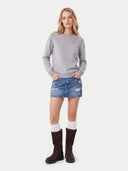 A woman modeling a light gray crewneck sweater, a distressed blue denim mini skirt, white socks, and dark brown boots, standing against a solid off-white background.