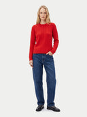 A woman stands against a light gray studio background wearing a bright red cable-knit sweater, blue jeans, and black leather shoes.