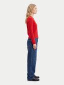 A woman shown in side profile wearing a bright red knit sweater, blue jeans, and black shoes against a plain light gray studio background.