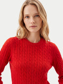 A blonde woman wearing a bright red cable-knit crewneck sweater, shown from the chest up against a light gray studio background.