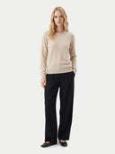 A woman stands against a light gray studio background wearing a light beige crew neck sweater, black wide-leg trousers, and black flats.