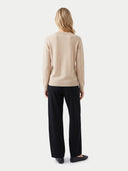 A woman viewed from the back wearing a light beige knit sweater and black wide-leg trousers against a plain light gray studio background.