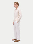 A man in a cream long-sleeve knit polo shirt and white trousers, standing in profile against a light gray studio background.
