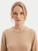 A close-up shot of a woman with blonde hair wearing a light beige crew-neck knit sweater against a plain light gray background.