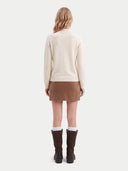 A woman is shown from the back wearing a cream mock-neck knit sweater, a brown mini skirt, white socks, and dark brown knee-high boots against a light gray studio background.
