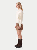 A woman stands in profile wearing a cream mock-neck knit sweater, a brown mini skirt, and dark brown mid-calf boots against a plain light gray studio background.