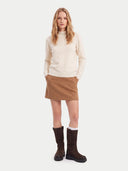 A woman stands against a light gray studio background wearing a cream mock-neck knit sweater, a brown mini skirt, white socks, and dark brown boots.
