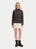A woman stands against a light gray studio background wearing a dark brown mock-neck knit sweater, a cream mini skirt, white socks, and black boots.