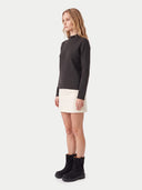 A woman stands in profile wearing a dark charcoal gray mock-neck knit sweater, a cream-colored mini skirt, and black ankle boots against a light gray studio background.