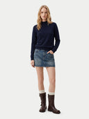 A woman stands against a light gray studio background wearing a dark navy mock-neck knit sweater, a blue denim mini skirt, cream socks, and dark brown leather boots.