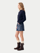 A woman stands in profile wearing a dark navy long-sleeve knit top, a blue denim mini skirt, cream ribbed socks, and dark brown ankle boots, against a plain light gray studio background.