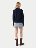A woman is shown from the back wearing a navy blue mock-neck knit sweater, a light blue denim mini skirt, cream socks, and dark brown leather boots, set against a plain light gray studio background.