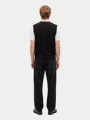 A man viewed from behind wearing a black sleeveless knit sweater vest over a white short-sleeve t-shirt, paired with black jeans and black shoes, against a light gray studio background.