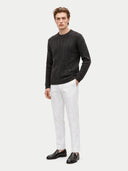 A man modeling a dark charcoal gray textured knit sweater, white trousers, and black leather loafers against a light gray studio background.