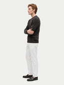 A man in profile wearing a dark charcoal textured knit sweater, white trousers, and black loafers, standing against a light gray studio background.