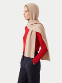 A woman in profile wearing a beige ribbed hijab draped over a red knit sweater and dark navy trousers, photographed against a light gray studio background.
