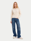 A woman modeling a cream cable-knit crewneck sweater, blue wide-leg jeans, and off-white sneakers against a light gray studio background.