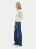 A woman in a side profile wearing a cream cable-knit sweater, blue wide-leg jeans, and white sneakers, photographed against a light gray studio background.