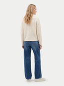 A woman shown from the back wearing a cream cable-knit sweater, blue wide-leg jeans, and white sneakers against a light gray studio background.