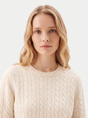 A blonde woman wearing a cream-colored cable-knit crewneck sweater, photographed against a clean, light gray studio background.