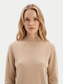 A woman with blonde hair wearing a beige knit mock neck sweater, photographed against a clean light gray background.