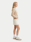 A woman stands in profile wearing a light beige knit sweater, an off-white mini skirt, white socks, and cream sneakers against a plain light gray studio background.