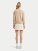 A woman viewed from the back wearing a beige mock-neck knit sweater, a cream mini skirt, white socks, and light-colored sneakers against a light gray studio background.