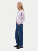 A woman stands in profile wearing a light lavender knit button-front cardigan over a white top, blue wide-leg jeans, and black loafers, photographed against a plain light gray studio background.