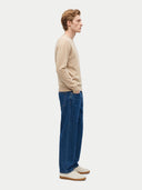 A man in profile wearing a beige knit sweater, blue jeans, and cream sneakers, standing against a light gray studio background.