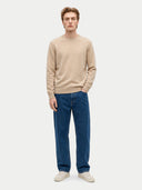 A male model stands against a light gray studio background wearing a beige crewneck sweater, dark blue jeans, and off-white sneakers.