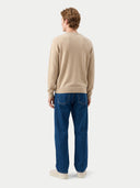 A man viewed from behind wearing a beige knit sweater, blue jeans, and off-white sneakers against a light gray studio background.