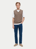 A male model stands against a light gray studio background wearing a light brown textured knit sweater vest over a white t-shirt, paired with blue jeans and beige flats.
