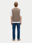 A man viewed from the back wearing a taupe cable-knit sweater vest over a white t-shirt, blue jeans, and tan shoes, against a light gray studio background.