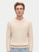 A man wearing a cream-colored cable-knit crewneck sweater and light blue jeans, standing against a solid light gray background.