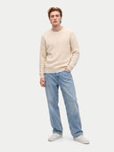 A man modeling a cream cable-knit crewneck sweater, light-wash blue jeans, and white sneakers against a light gray studio background.