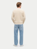 A man viewed from behind wearing a cream cable-knit sweater, light blue jeans, and white sneakers, standing against a light gray studio background.