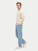 A man wearing a cream cable-knit sweater, light blue jeans, and white sneakers, standing in profile against a light gray studio background.