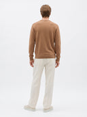 A man viewed from behind wearing a light brown crewneck sweater, off-white trousers, and light-colored shoes, standing against a plain white studio background.