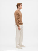 A man in profile wearing a tan crewneck sweater, off-white trousers, and off-white sneakers, standing against a plain white studio background.