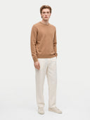 A man modeling a light brown crewneck sweater paired with off-white trousers and light beige sneakers, standing against a plain light gray studio background.