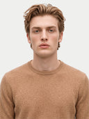A close-up portrait of a young man with light brown hair wearing a warm tan crew-neck knit sweater against a light gray background.