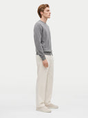 A man in profile wearing a gray long-sleeve knit sweater, off-white trousers, and light beige sneakers, standing against a plain light gray studio background.