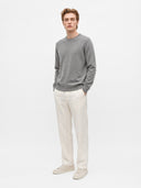 A man modeling a gray crewneck sweater, off-white trousers, and beige sneakers against a plain white studio background.