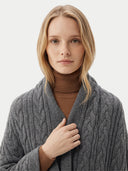 A blonde woman wearing a chunky gray cable-knit cardigan over a brown turtleneck sweater, photographed against a clean light gray studio background.