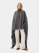 A woman stands against a light gray studio background wearing a large charcoal gray cable-knit poncho over a brown turtleneck, cream wide-leg pants, and brown slip-on shoes.