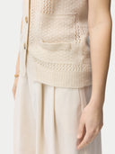 A woman wearing a beige sleeveless cashmere vest with a round neckline and button closures, paired with a white skirt. #color_off-white