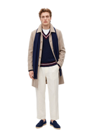 Man in a taupe single-breasted cashmere coat over a navy cable-knit cricket sweater, with a navy fringed scarf, off-white trousers and navy loafers